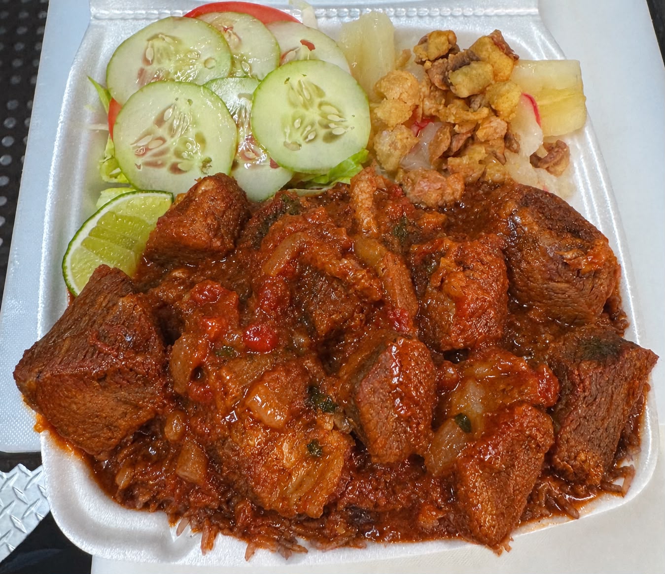 Stewed lamb in rich reddish sauce with congrí rice, yuca, chicharrones and salad in a takeout container